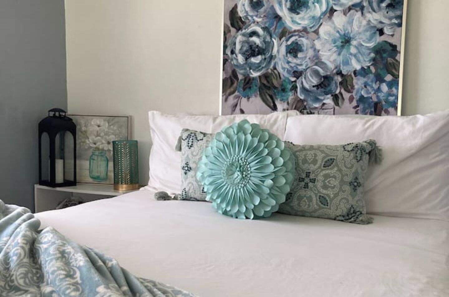 A neatly made bed with a large decorative pillow, a bedside table with a lantern, and a floral painting on the wall.