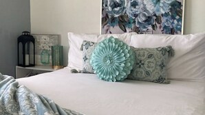 Un lit bien fait, agrémenté d’un grand coussin décoratif, une table de chevet avec une lanterne et un tableau floral accroché au mur.