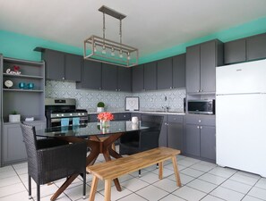 A modern kitchen with dark grey cabinets, a white refrigerator, a microwave, a stove, and a dining area with a glass table and wicker chairs.