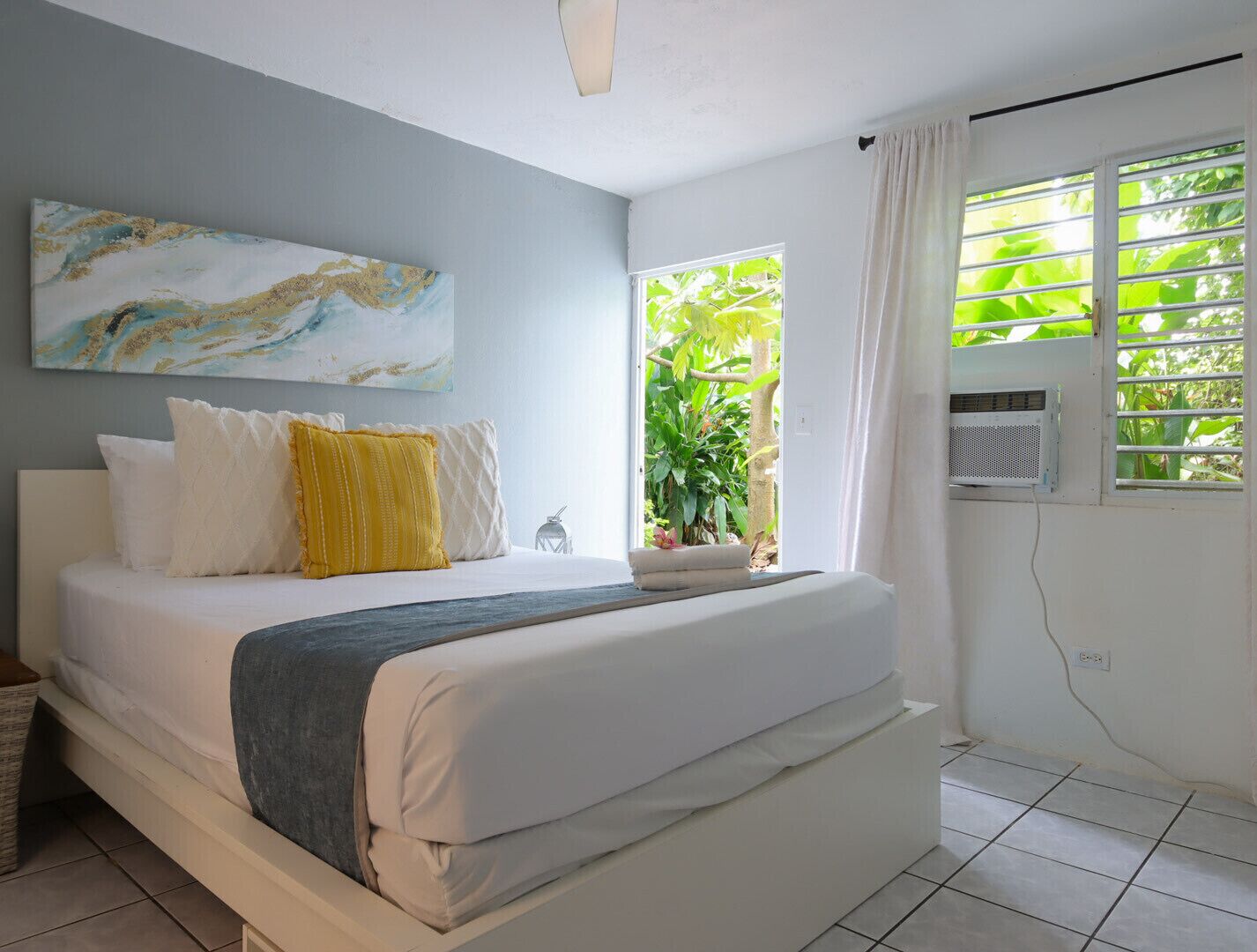 A bedroom with a bed, a window with curtains, a ceiling fan, and a wall-mounted air conditioner.