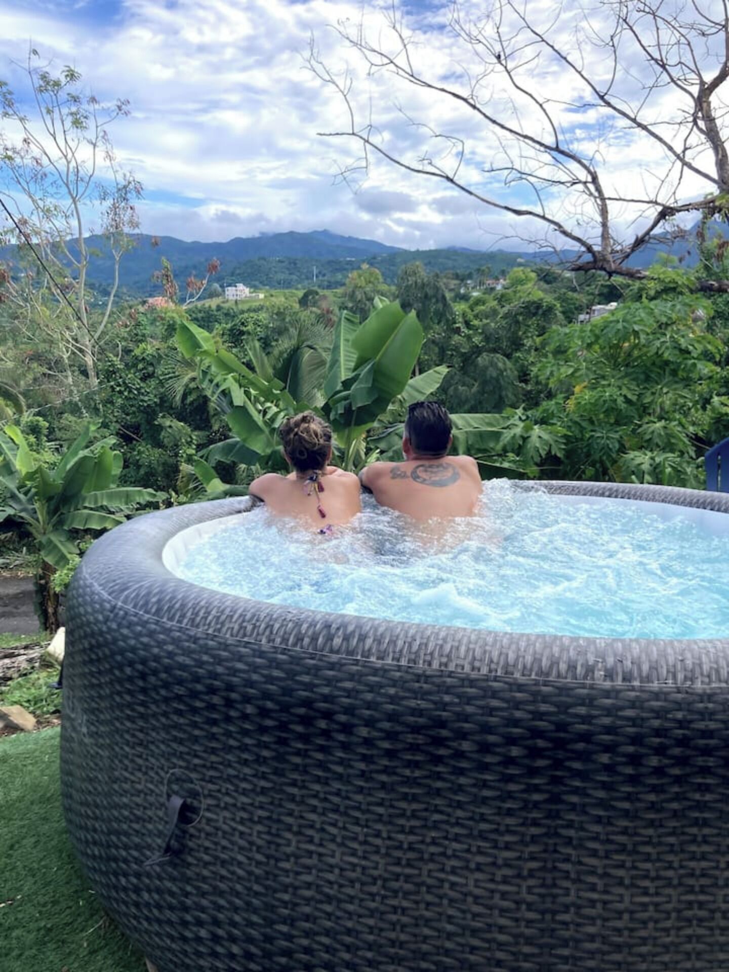 Ein Whirlpool mit zwei Personen darin, umgeben von üppigem Grün und Bergen im Hintergrund.