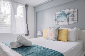 A neatly made bed with a blue patterned bedspread, a pair of rolled towels, and a wall with a sea turtle artwork. - Tropical Retreat with Spectacular Rooftop Rainforest & Ocean Views With Jacuzzi! (Luquillo)