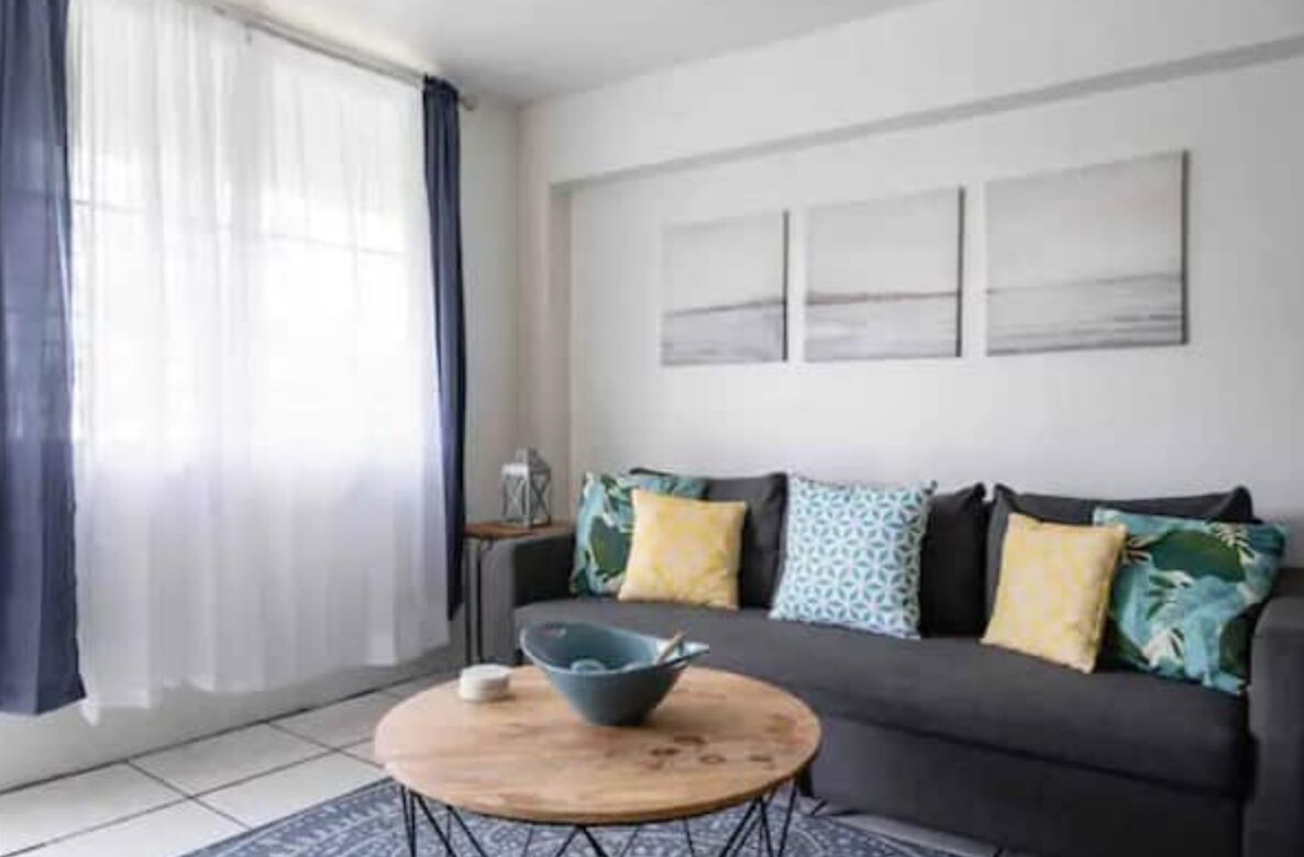 Una sala moderna con un sofá gris, una mesa de centro redonda de madera y una ventana con cortinas translúcidas.