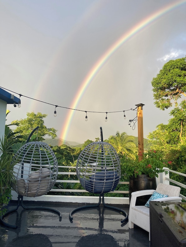 A rooftop terrace with two hanging egg chairs, a string of lights, and a view of a rainbow.