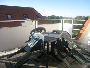 Ein Balkon mit Tisch und vier Stühlen, im Hintergrund ein Gebäude mit rotem Dach und ein weißer Zaun.