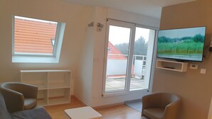 Ein modernes Wohnzimmer mit einer Couch, Sesseln, einem kleinen Tisch, einem an der Wand befestigten Fernseher und einer Schiebetür zu einem Außenbereich.