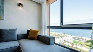 A modern living room with a grey sofa, a yellow pillow, and a large window overlooking a beach and cityscape. - Atlantic View Appart'Hotel (Casablanca)