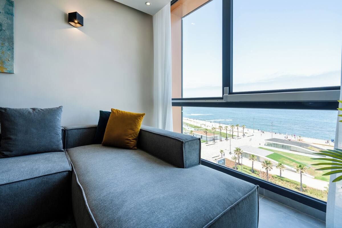 Ein modernes Wohnzimmer mit grauem Sofa, einem gelben Kissen und einem großen Fenster mit Blick auf Strand und Stadtlandschaft.