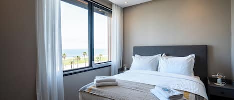 A hotel room with a large bed, a nightstand, a window with a view of the beach, and a ceiling light.