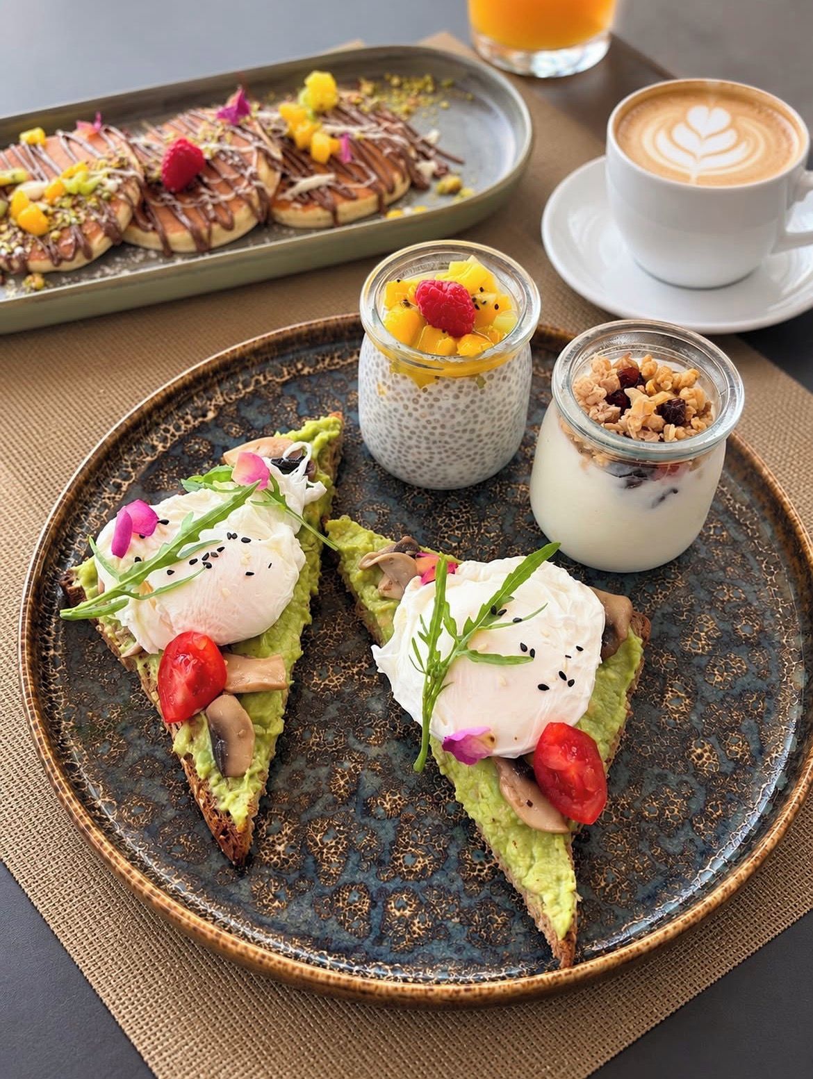 A plate with avocado toast topped with poached eggs, a small jar of chia pudding, and a glass of orange juice.