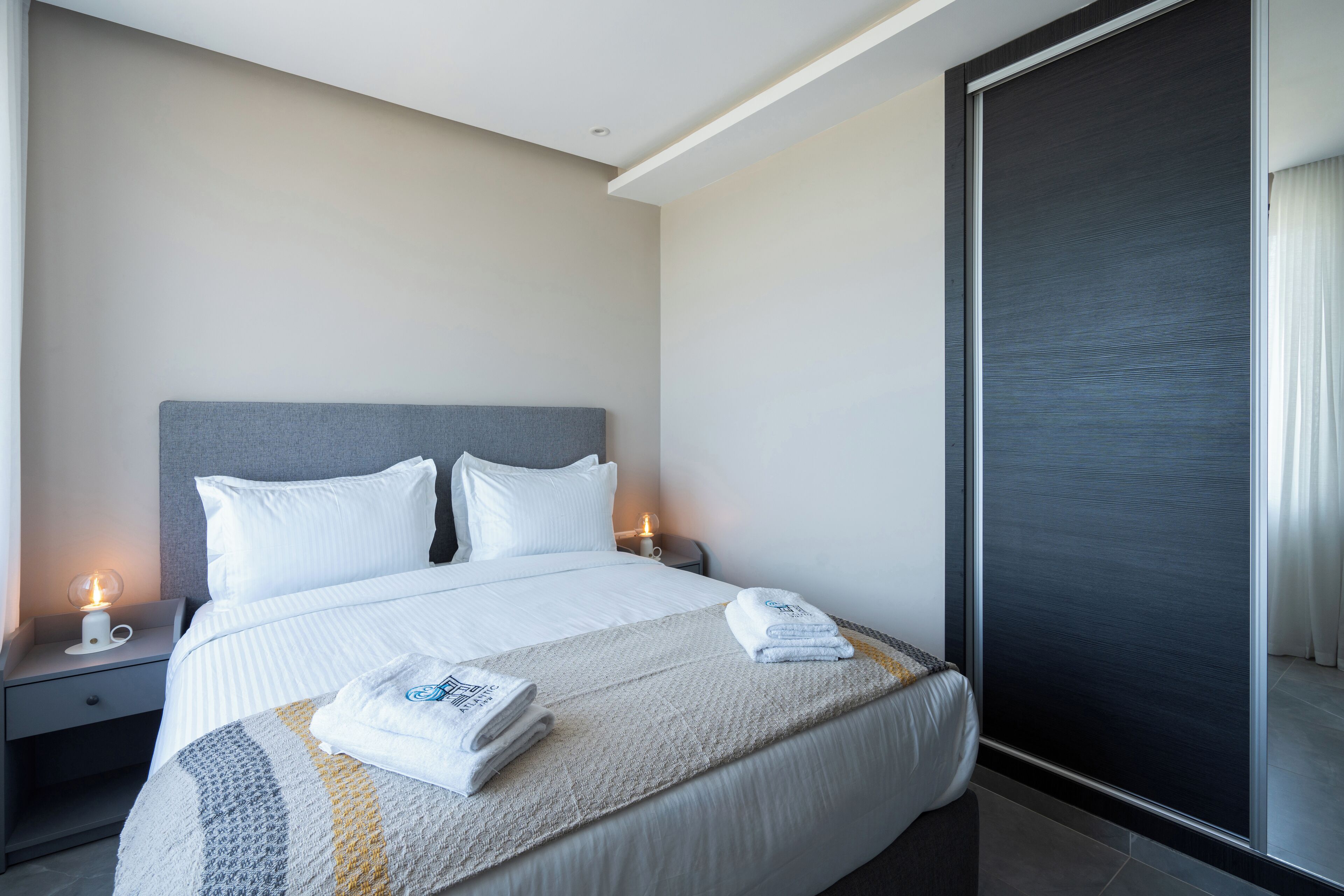 A neatly made bed with white linens, a grey headboard, and a bedside table with a lamp and a cup.