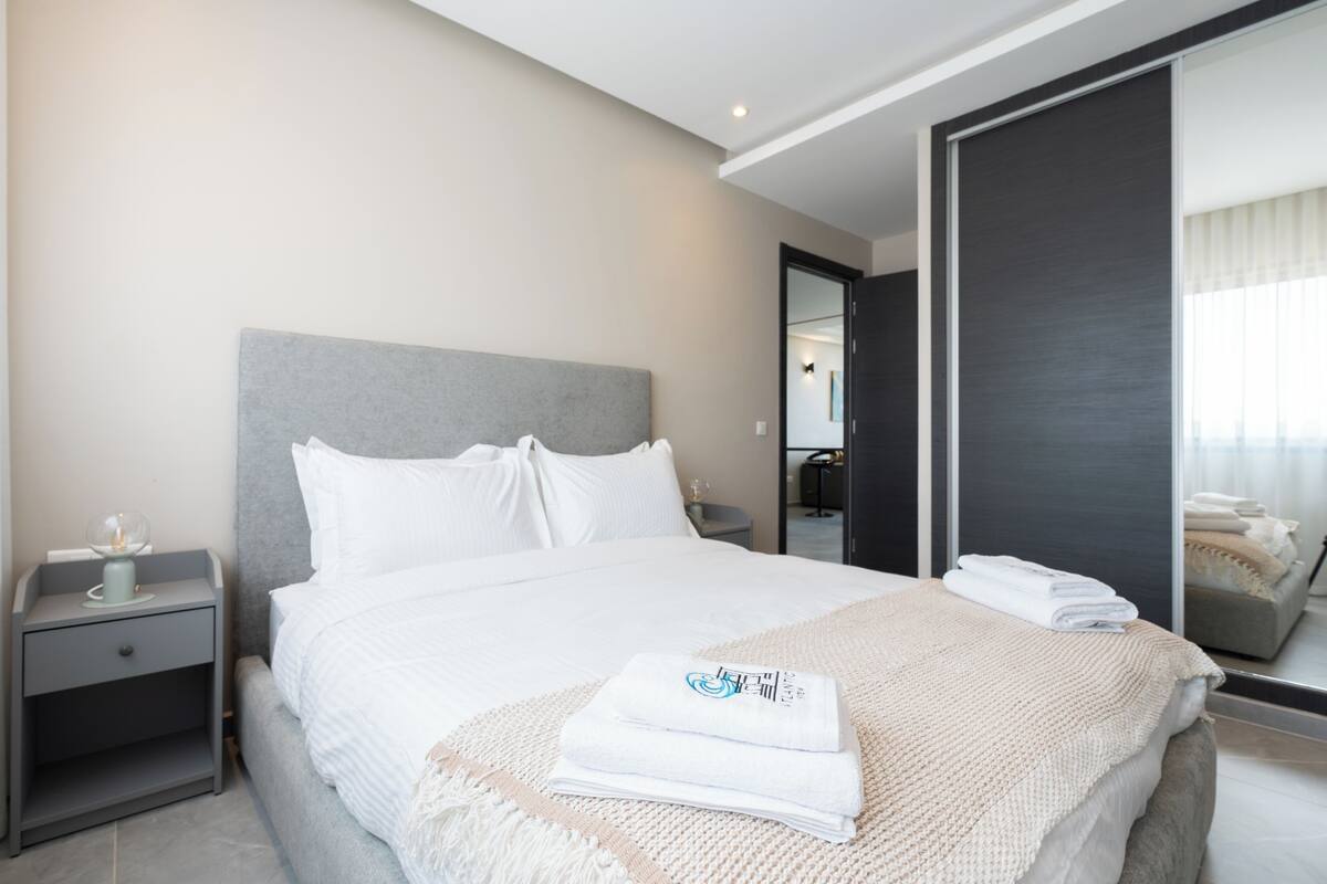 Ein modernes Hotelzimmer mit einem großen Bett, einem Nachttisch mit Lampe und einem Spiegel, der den Raum reflektiert.