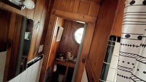 A narrow wooden hallway with a sink, mirror, and towel rack on the left. A door leads to a small kitchen area with a window.