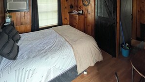 A wooden cabin room with a bed, a television, a window with curtains, a wall-mounted heater, and a sliding door.