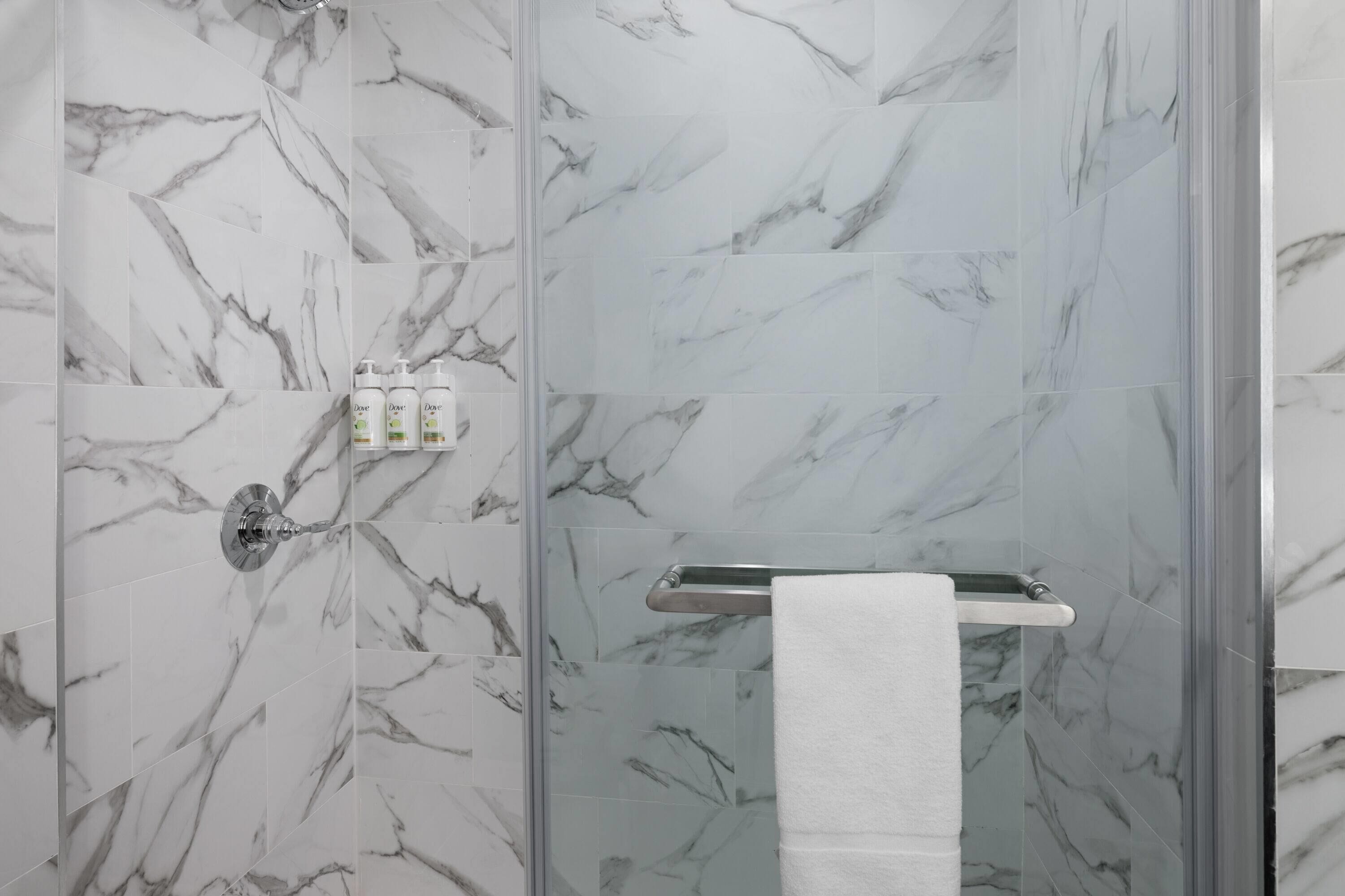 Une salle de bain carrelée de marbre, dotée d’une douche en verre, d’un porte-serviettes et d’un espace mural pour les articles de toilette.