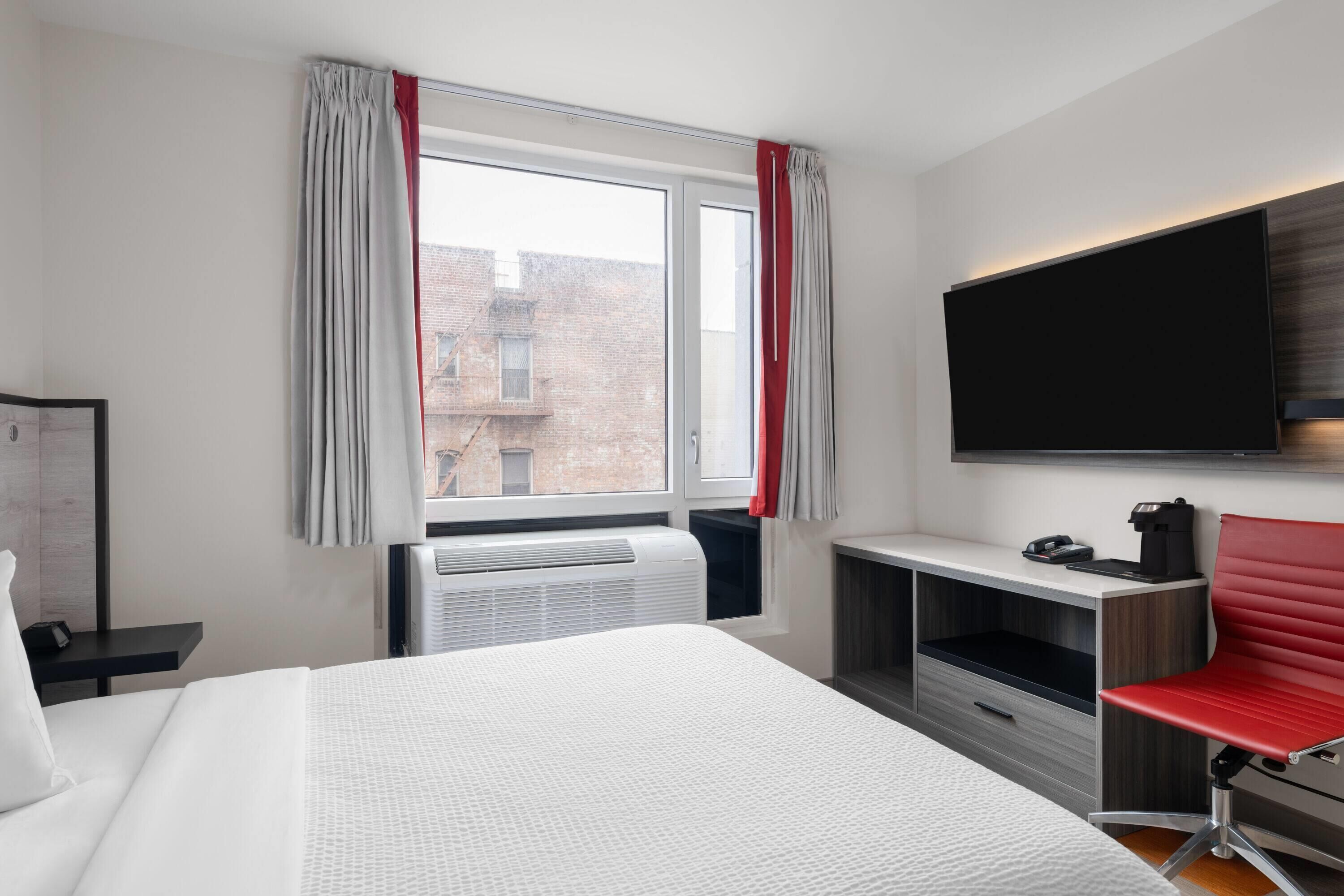 A hotel room with a large bed, a desk with a flat-screen TV, a red chair, and a window with curtains.