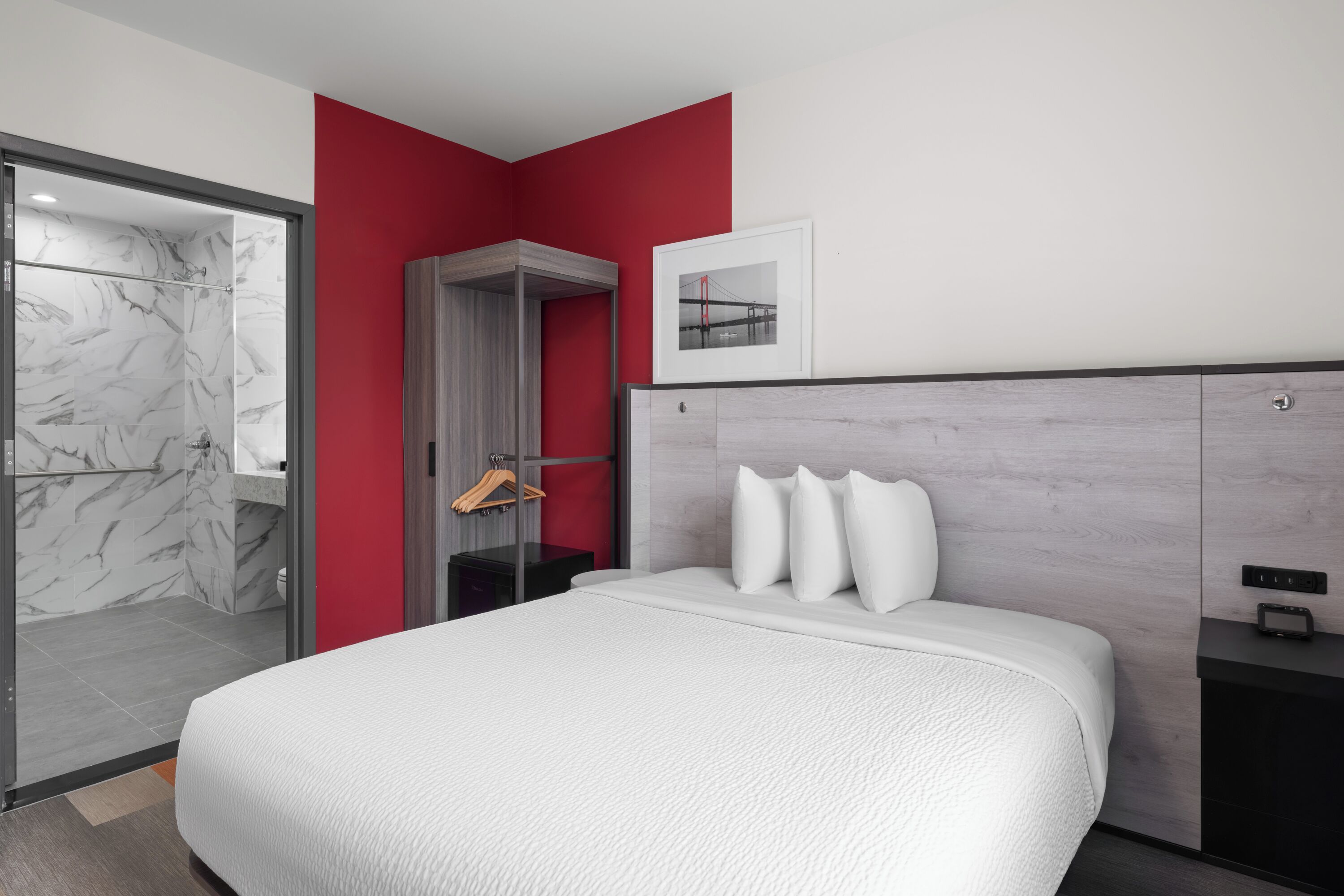 Une chambre d’hôtel moderne avec un lit, un mur d’accent rouge, un tableau encadré et un espace douche.