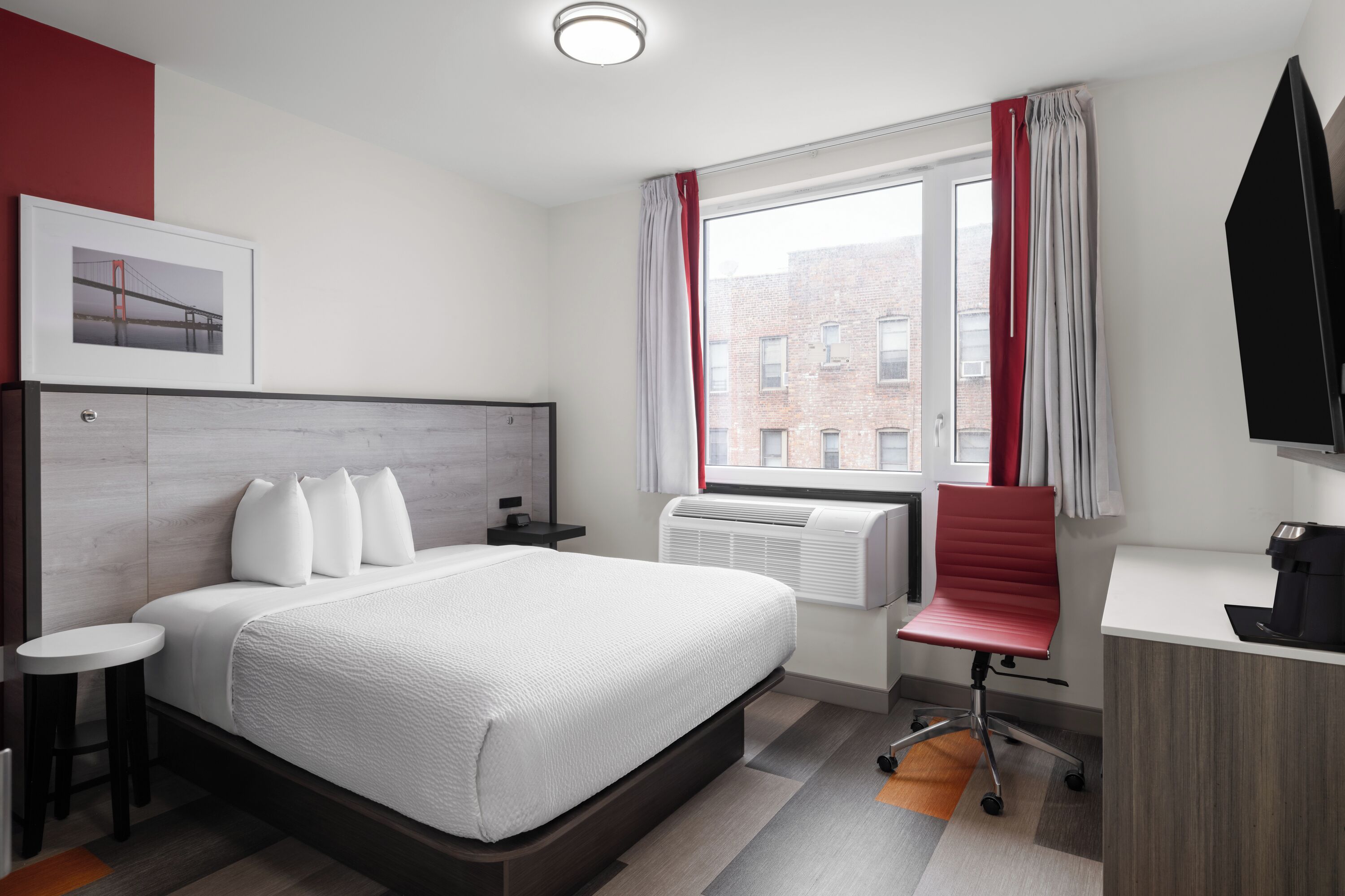 Une chambre d’hôtel comprenant un lit, un bureau, une chaise, une machine à café, une fenêtre avec des rideaux, un tableau au mur et un téléviseur.