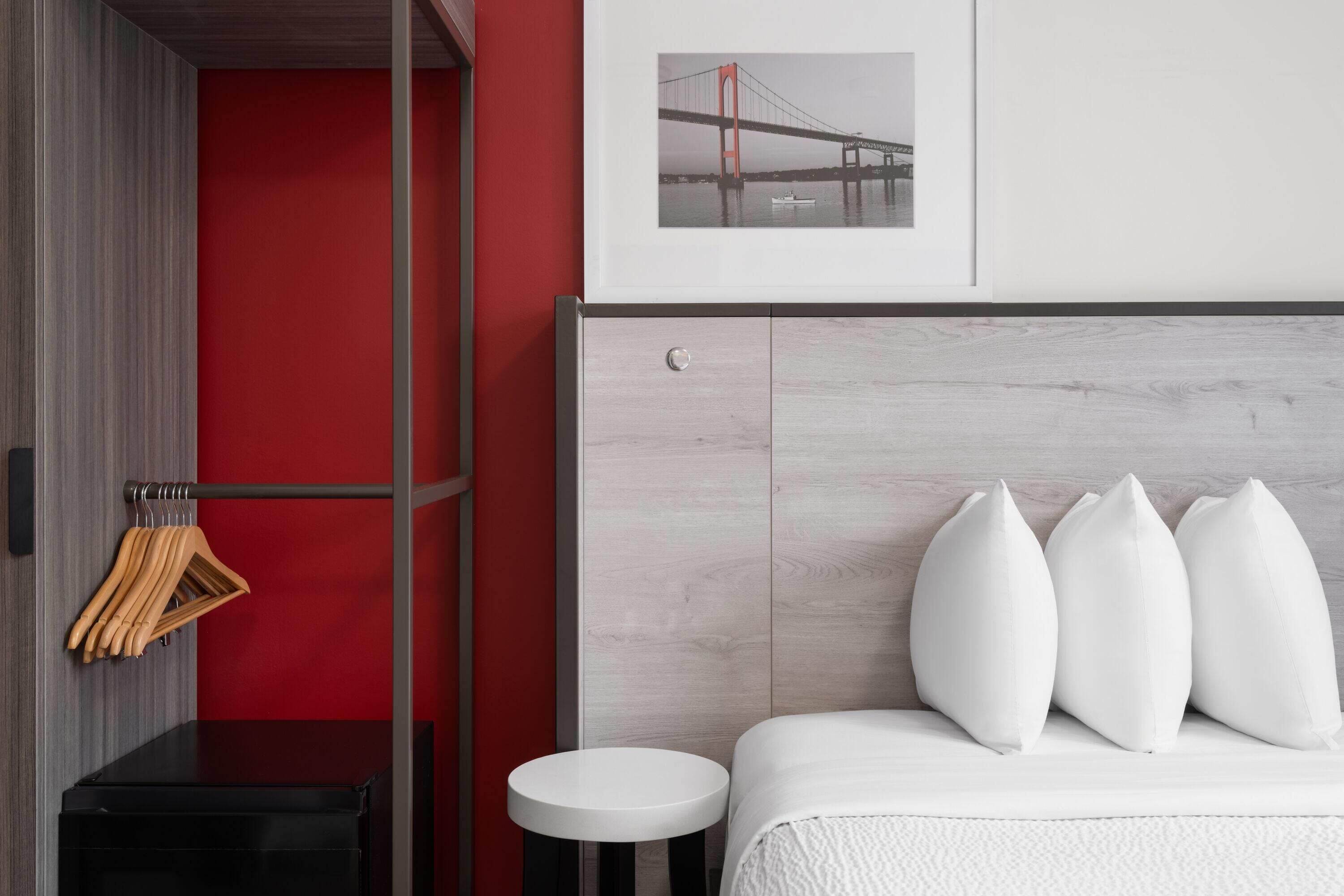 Quarto de hotel moderno com parede de destaque vermelha, uma imagem emoldurada de uma ponte, cama com almofadas brancas, mesa redonda pequena e mesa de cabeceira preta.