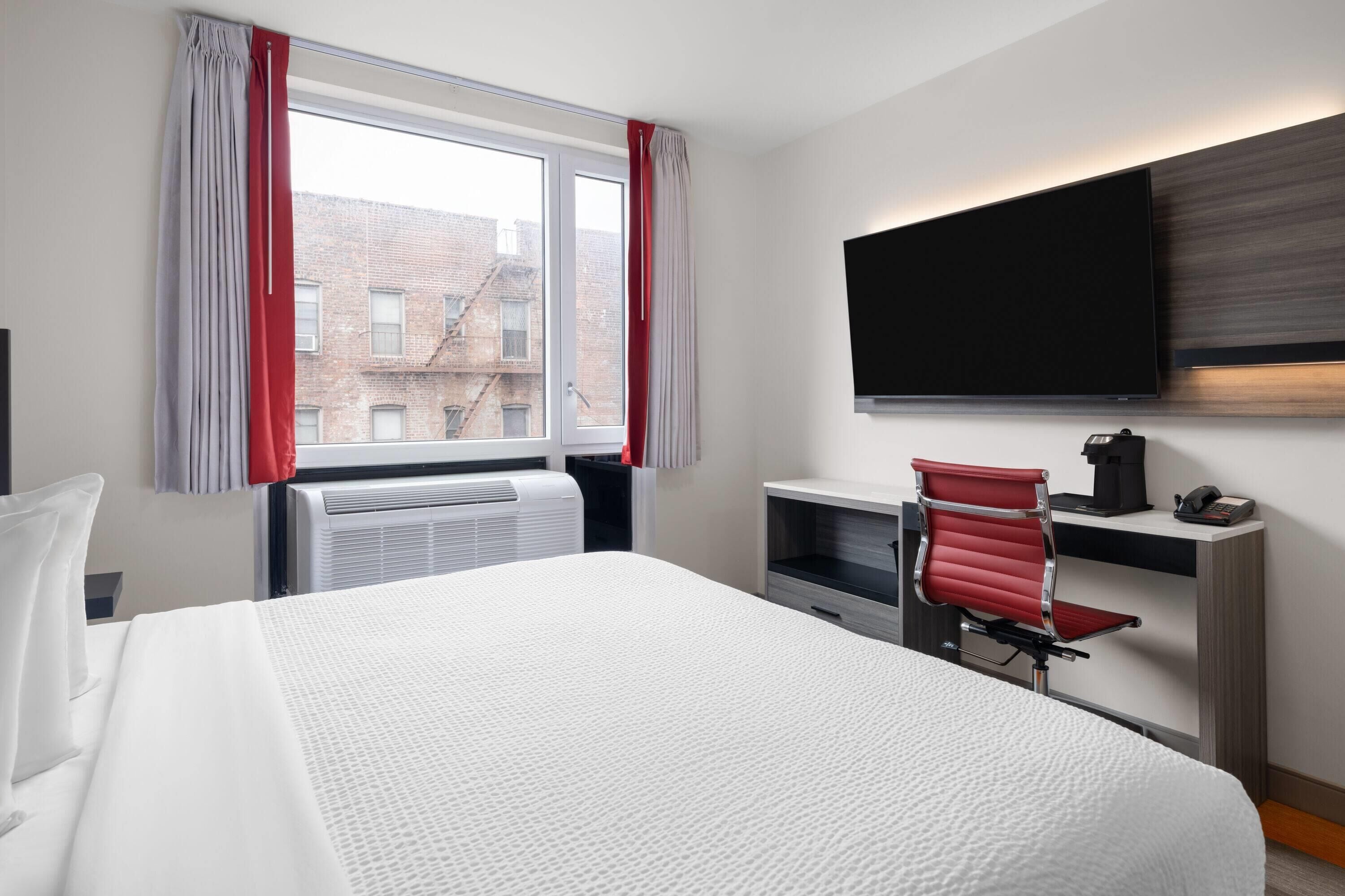 A hotel room with a large bed, a desk with a chair, a flat-screen TV mounted on the wall, and a window with curtains.