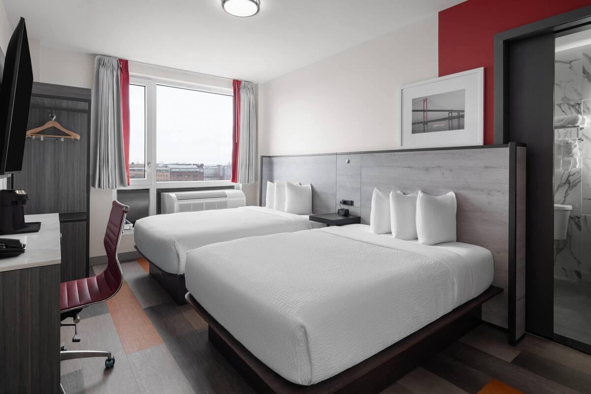 Ein Hotelzimmer mit zwei Betten, einem Schreibtisch mit Stuhl, einem Fernseher und einem Fenster mit Vorhängen.