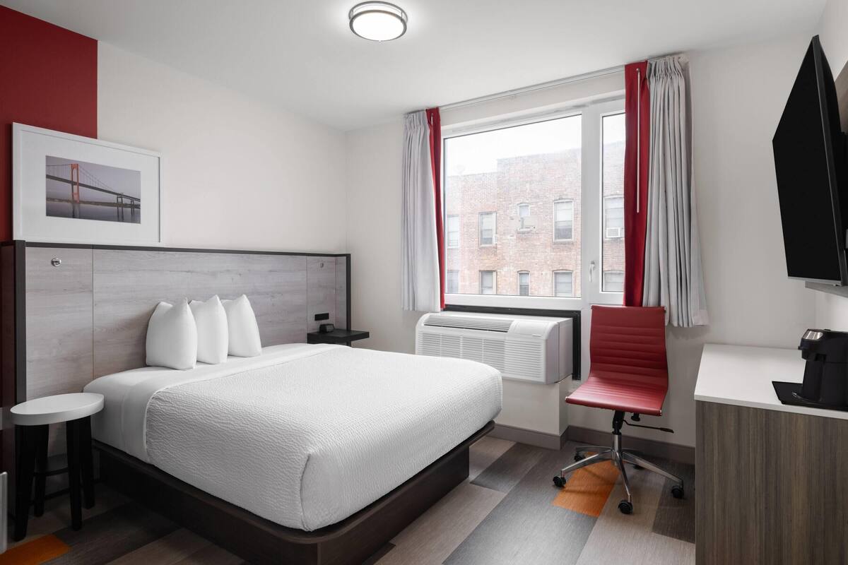 Ein modernes Hotelzimmer mit einem großen Bett, einem Schreibtisch, einem Sessel, einer Kaffeemaschine und einem Fenster mit Vorhängen.