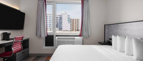 A hotel room with a large bed, a desk with a chair, a flat-screen TV, and a view of city buildings through the window.