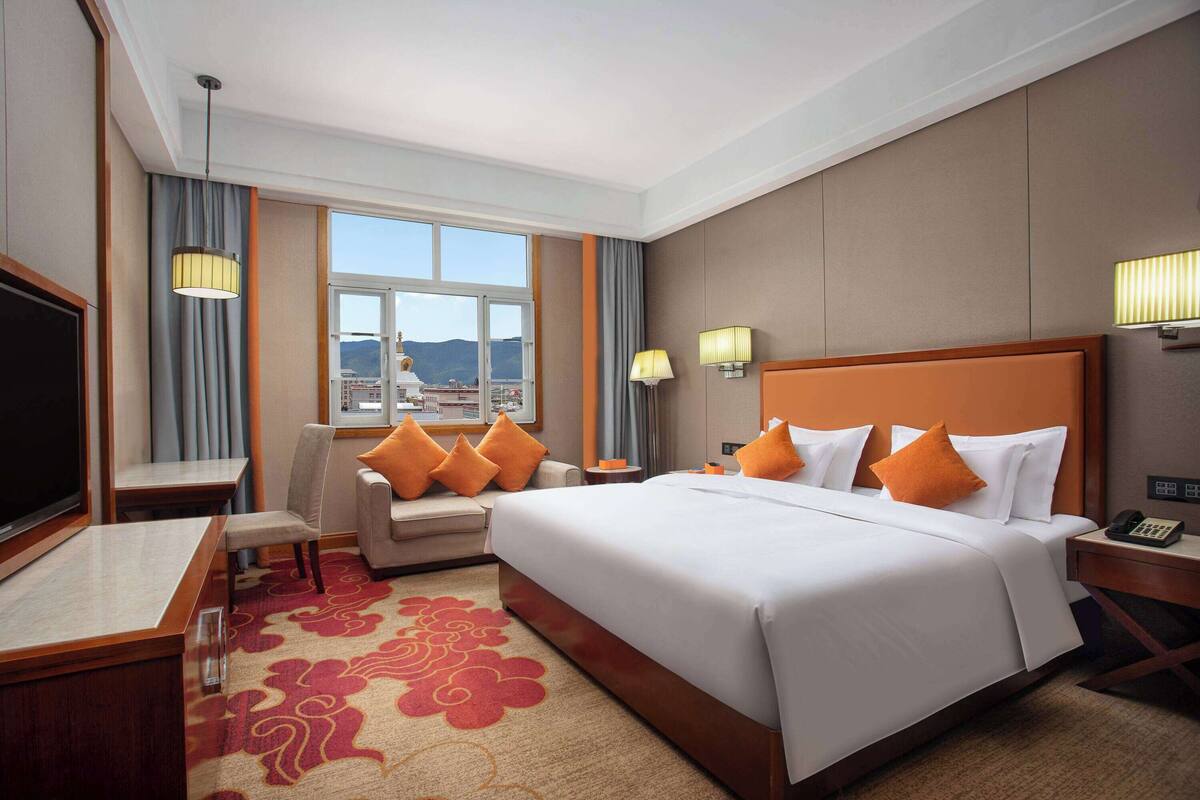 Ein Hotelzimmer mit einem großen Bett, einem Schreibtisch, einem Sessel und einem Fernseher. Ein Fenster bietet einen Blick auf die Berge.
