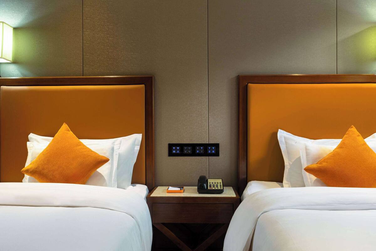 Ein Hotelzimmer mit zwei Betten, jedes mit einem hölzernen Kopfteil und einem einzelnen orangefarbenen Kissen. Ein Nachttisch mit Telefon und Notizblock ist vorhanden.