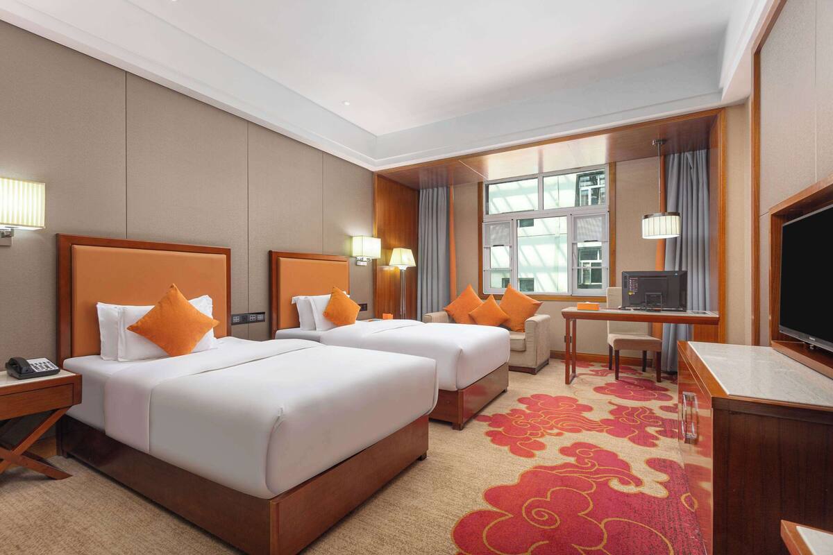 Ein Hotelzimmer mit zwei Betten, einem Schreibtisch, einem Fernseher und einem Fenster mit Vorhängen.