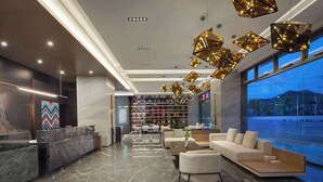 Un hall d’hôtel moderne doté d’un comptoir d’accueil, d’un espace salon et de grandes fenêtres offrant une vue sur le paysage urbain.