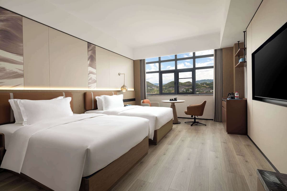 Ein Hotelzimmer mit zwei Betten, einem Schreibtisch, einem Sessel, einem Fernseher und einem Fenster mit Blick auf die Hügel.
