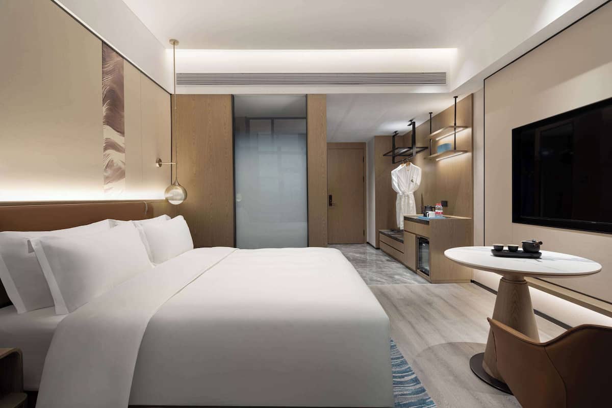 Ein modernes Hotelzimmer mit einem großen Bett, einem kleinen runden Tisch, einem Fernseher und einem Badezimmer mit Spiegel und Waschbecken.