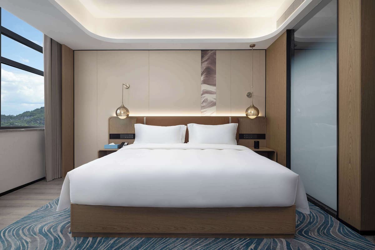 Ein modernes Hotelzimmer mit einem großen Bett, zwei Nachttischlampen, einem Nachttisch und Blick ins Freie.