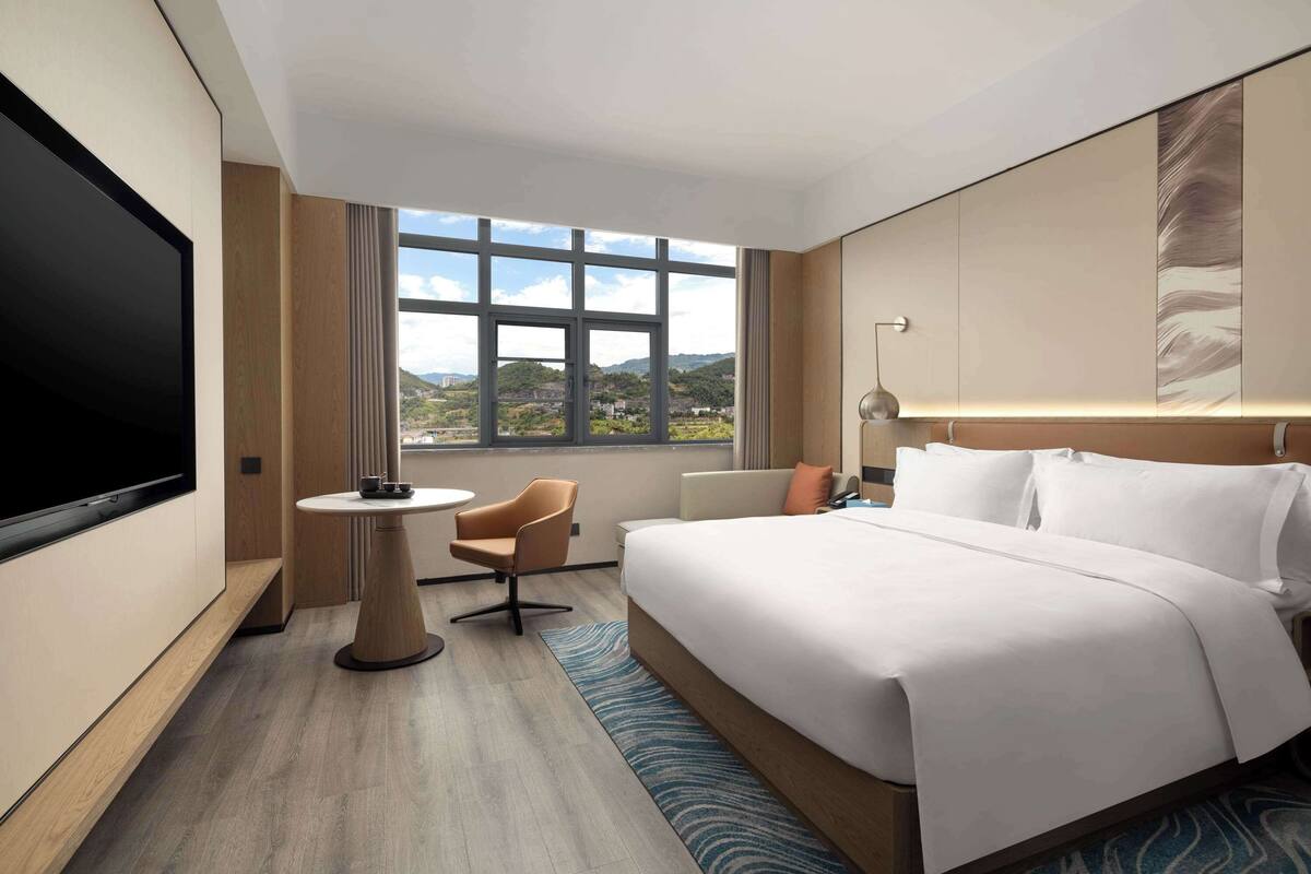 Ein modernes Hotelzimmer mit einem großen Bett, einem Flachbildfernseher, einem kleinen runden Tisch, einem Sessel und Blick ins Freie.