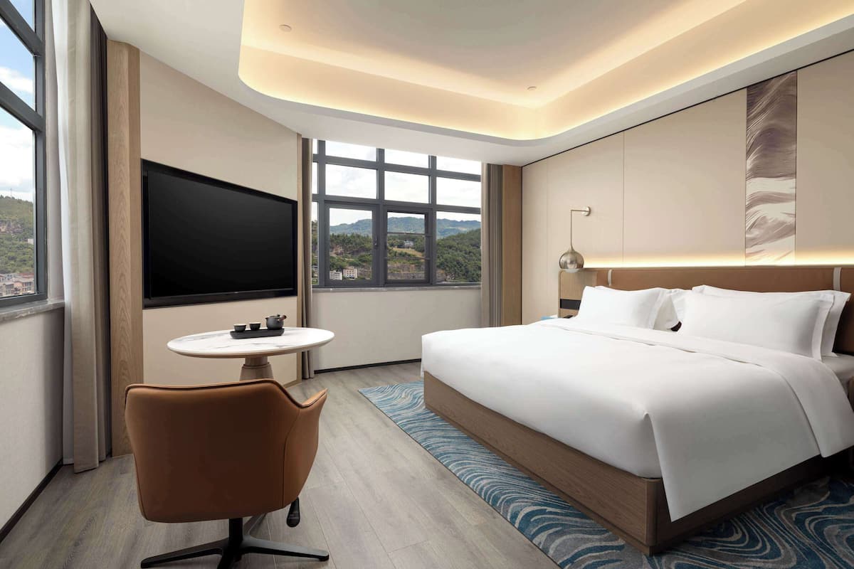 Ein modernes Hotelzimmer mit einem großen Bett, einem Flachbildfernseher, einem kleinen runden Tisch und einem Sessel.