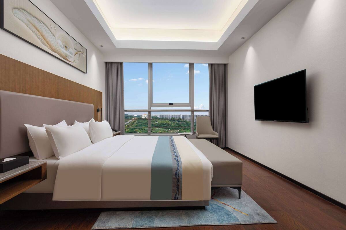 A modern hotel room with a large bed, a flat-screen TV, and a view of a cityscape through large windows.