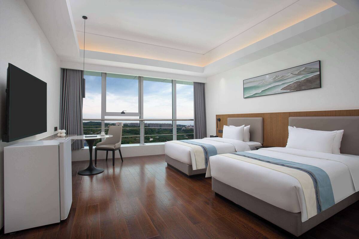 A hotel room with two beds, a desk, a chair, a TV, and a large window offering a city view.