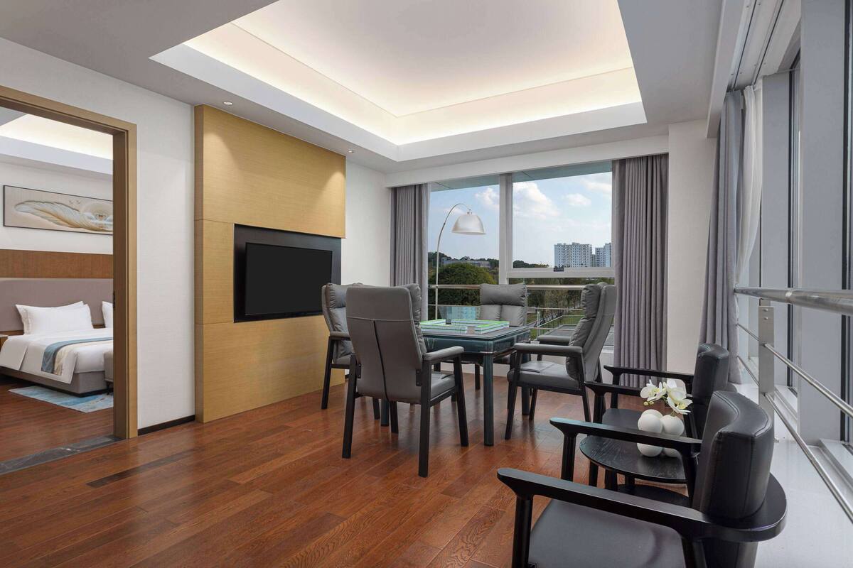 A modern hotel room with a flat-screen TV, a dining area with a table and chairs, a bed with a headboard, and a large window offering a city view.