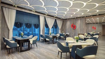 Uma zona de refeições moderna com mesas e cadeiras, amplas janelas com cortinas e um teto decorativo.