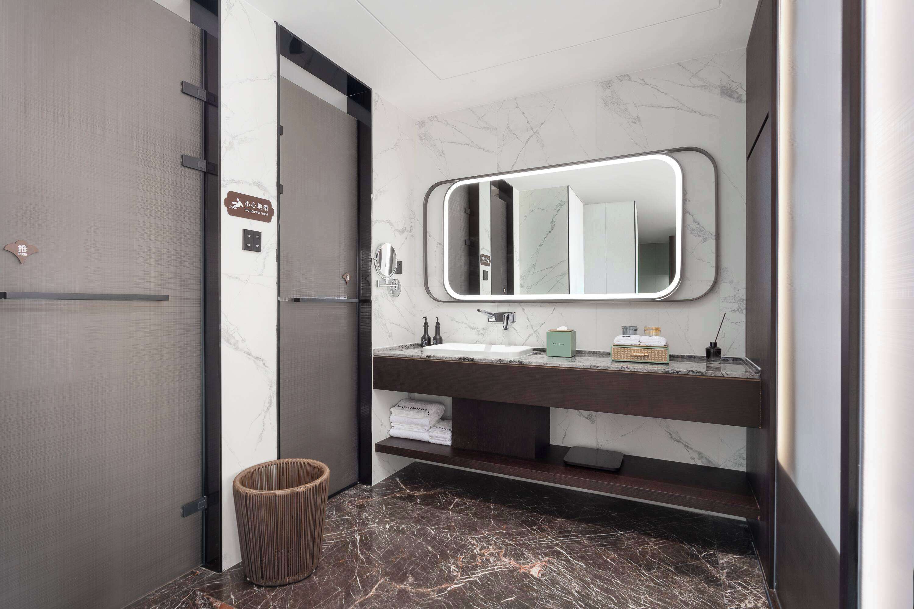 Un baño moderno con piso de mármol, tocador oscuro, espejo grande y un cesto de basura.