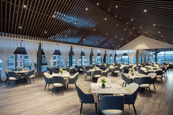 A spacious dining area with multiple tables and chairs, large windows with curtains, and a high ceiling with a slatted design. - Wyndham Grand Xishuangbanna Resort (Xishuangbanna Dai)
