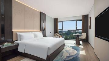 A modern hotel room with a large bed, a desk, a chair, a TV, and a balcony with a city view.