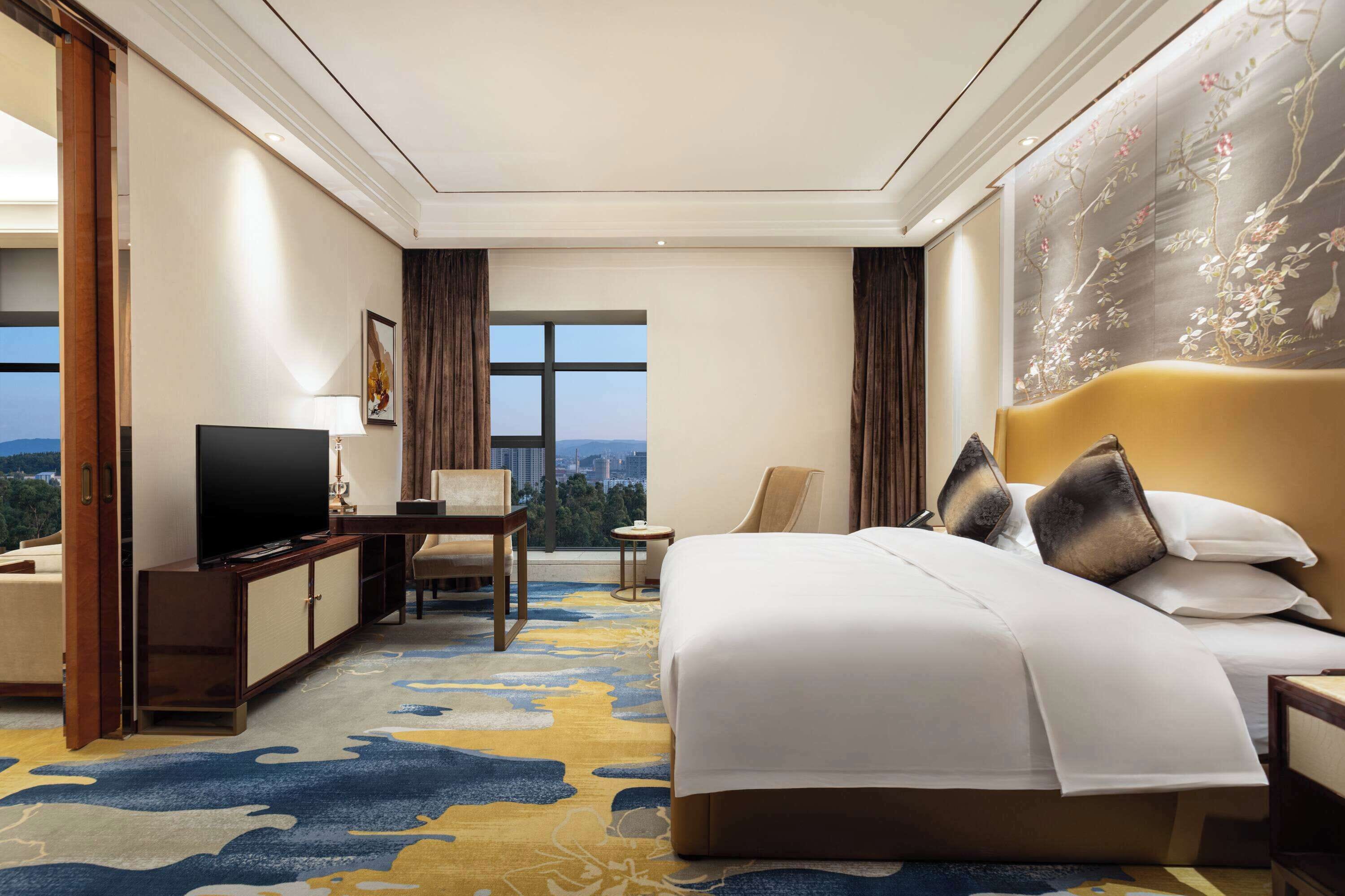 A hotel room with a large bed, a television, a desk, and a view of the city.