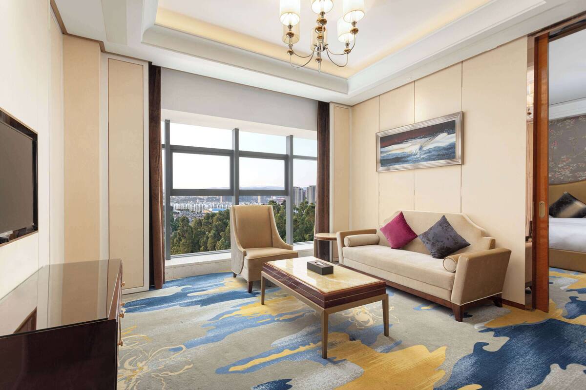Quarto de hotel com uma janela grande, um sofá, uma poltrona, uma mesa de centro e uma televisão.