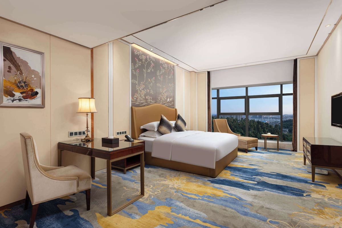 Quarto de hotel com uma cama grande, uma escrivaninha com uma luminária, uma cadeira, uma mesinha e uma televisão.