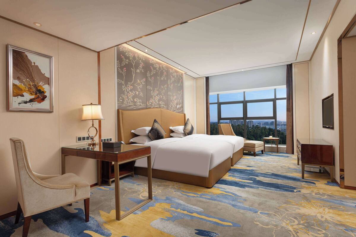 Quarto de hotel com uma cama grande, uma escrivaninha com abajur, uma cadeira, uma televisão e vista para a cidade.