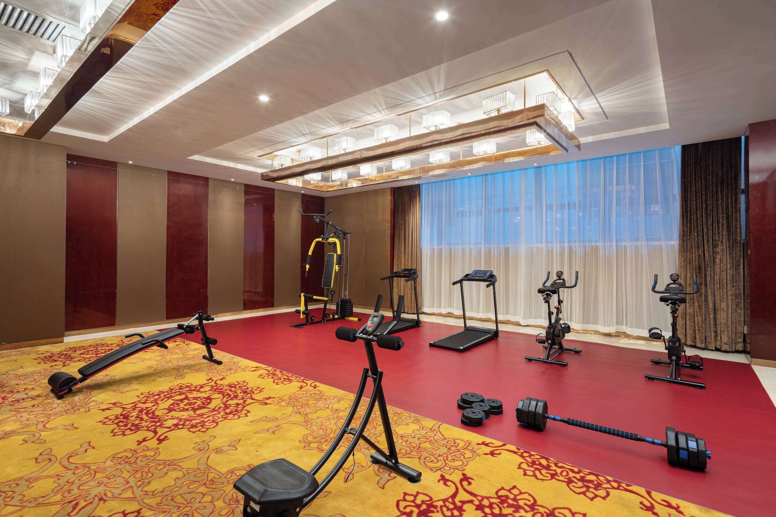 A hotel room with a red carpet, exercise equipment, and a large window with curtains.