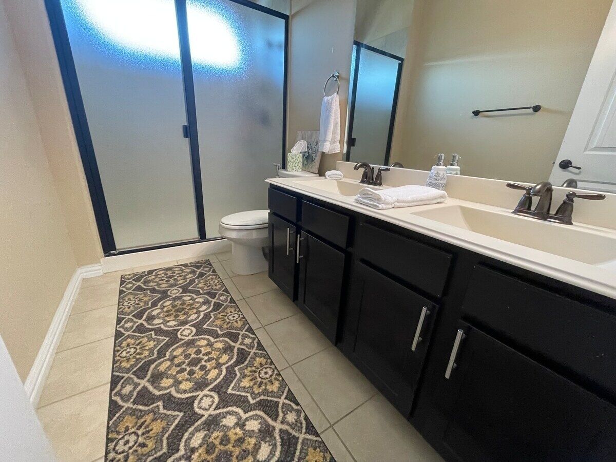 Une salle de bain avec un grand miroir, deux lavabos, des toilettes et un tapis à motifs.