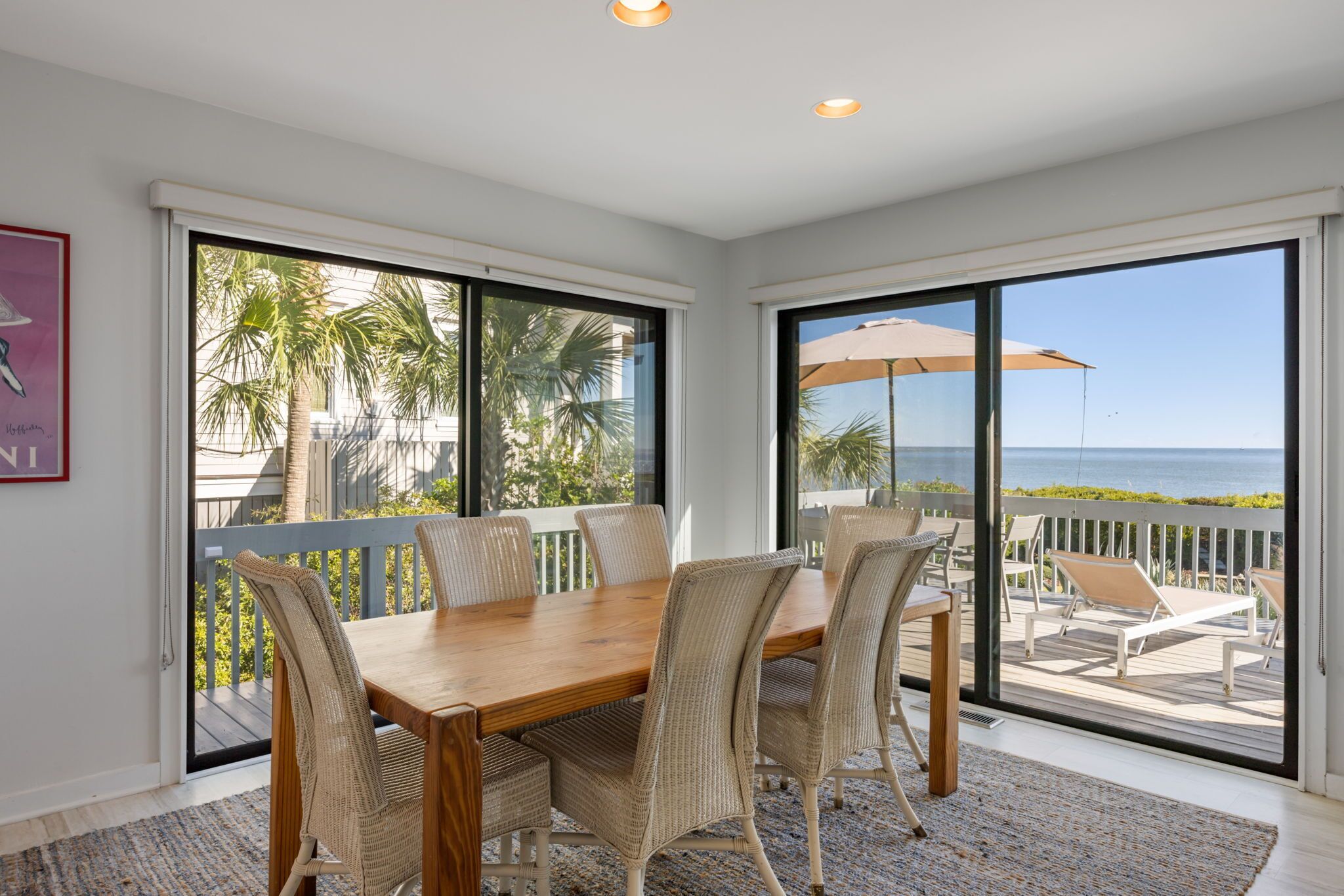 A dining area with a wooden table and wicker chairs, large sliding glass doors leading to a balcony with a view of the ocean.